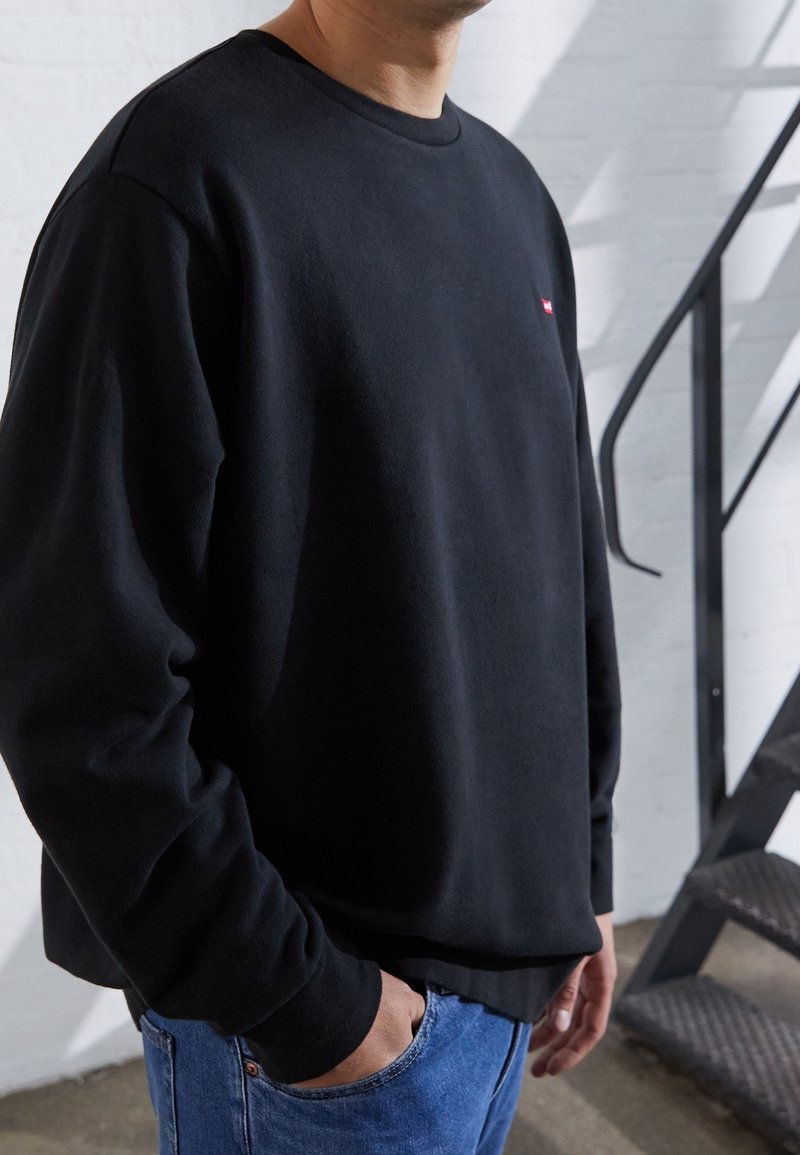 Sweat-shirt noir à col rond avec manches longues, fabriqué en tissu doux, présentant un petit logo rouge sur la poitrine et un ourlet côtelé.