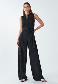 Ženska stoji v črnem brezrokavnem širokem kombinezonu z gumbi, pasom in belimi sandalami pred enostavno svetlo ozadje.