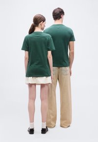 Δύο άτομα στέκονται πλάτη με πλάτη φορώντας σκουροπράσινα μπλουζάκια· το ένα φοράει μπεζ φούστα και μαύρα παπούτσια, το άλλο φαρδιά μπεζ παντελόνια και αθλητικά παπούτσια.