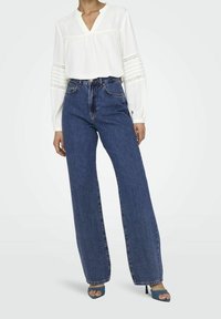 Valkoinen paita, jossa on pitkät hihat, V-pääntie ja pitsi-inserttit; yhdistetty korkeavyötäröisiin, leveisiin sinisiin farkkuihin ja korkokenkiin sinisillä remmeillä.