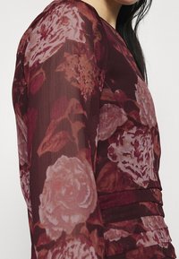 Blommig blus med långa ärmar, som har burgundy och rosa toner, genomskinlig textur och samlade detaljer på sidan.