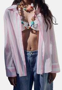 Camicia trasparente a righe rosa e bianche, indossata aperta sopra un top bikini bianco, abbinata a pantaloni denim blu a vita alta e una collana di perline colorate.
