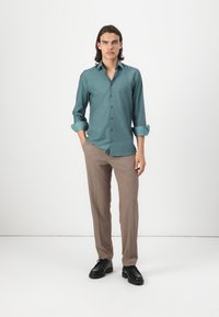Türkises Hemd mit Knopfverschluss und aufgerollten Ärmeln, kombiniert mit hellbraunen, taillierten Hosen und schwarzen Schuhen. Glattes Material und strukturierte Passform.