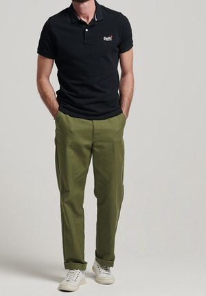 Hombre de pie con las manos en los bolsillos, lleva una camiseta polo negra, pantalones verde oliva con puños y zapatillas blancas sobre un fondo liso.