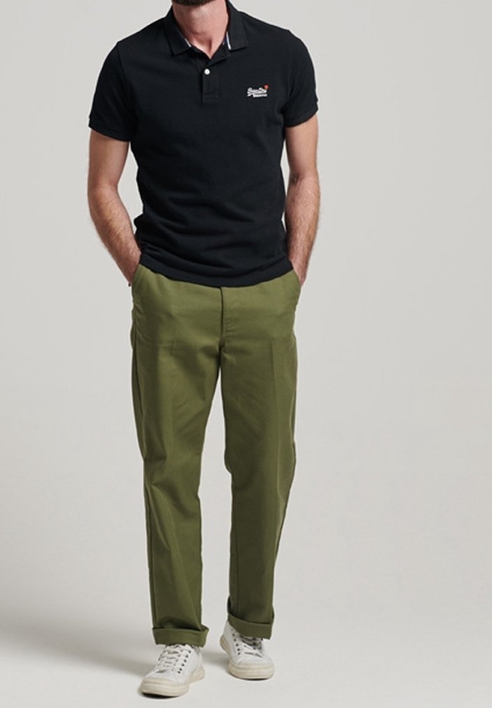 Un homme se tient debout, les mains dans les poches, portant un polo noir, un pantalon vert olive avec des revers et des baskets blanches sur un fond uni.