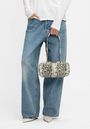 Persona che indossa jeans a gamba larga blu e scarpe bianche a punta, mentre tiene una piccola borsa rettangolare in pelle di serpente per le maniglie.
