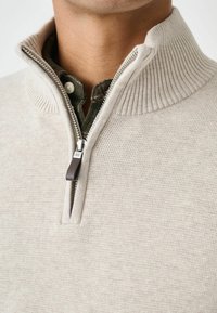 Gilet zippé beige clair avec un col côtelé, offrant une texture lisse et une fermeture éclair métallique avec une languette en cuir.