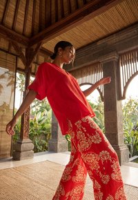 Mujer con camisa roja suelta y pantalones anchos de flores rojas, de pie dentro de un pabellón de madera con pilares tallados y plantas tropicales en el exterior.
