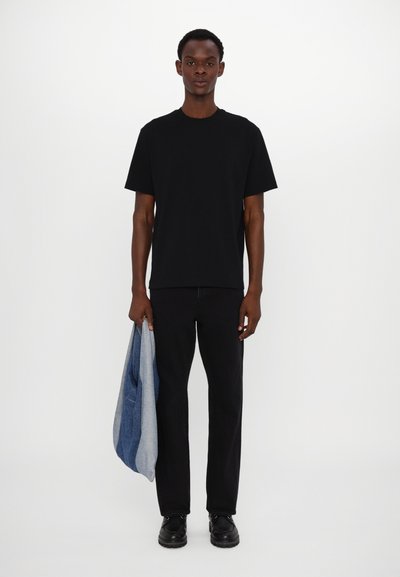 Tricou din bumbac negru cu mâneci scurte, asortat cu pantaloni negri și pantofi negri. O jachetă din denim albastru este aruncată pe brațul stâng.
