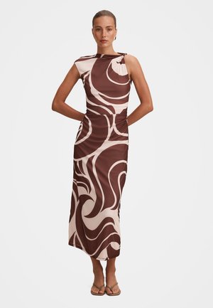 Vrouw met een mouwloze midi-jurk met bruin- en beige abstracte draaipatronen, staand met haar handen achter haar rug en sandalen aan.