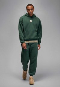 Grüner Hoodie mit einer Vordertasche und kleinem Logo, kombiniert mit passenden Jogginghosen. Das Material wirkt weich, mit lockerem Schnitt und gerippten Bündchen.