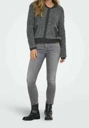Frau trägt eine graue, flauschige Strickjacke, hellgraue Skinny-Jeans und schwarze Stiefeletten mit Schnallendetails, steht vor einem schlichten Hintergrund.