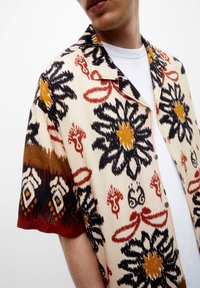 Chemise à manches courtes en tissu léger avec un fond crème, présentant des motifs floraux vibrants en noir, rouge, orange et marron.