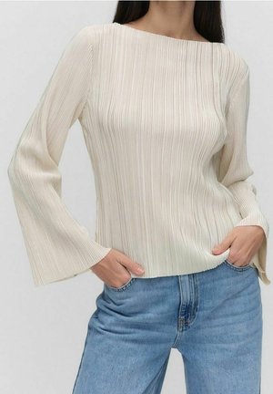 Vrouw draagt een crèmekleurige, geribbelde blouse met lange mouwen en brede manchetten, gecombineerd met lichtblauwe spijkerbroek, handen in de zakken.