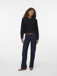 Zwarte gebreide trui, donkerblauwe wijde jeans, brown riem. Model staat in een neutrale achtergrond, draagt tan flats. Krullend haar.