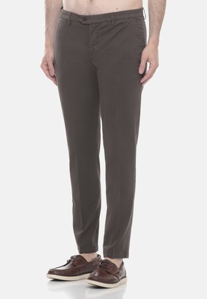 Uomo con pantaloni slim-fit marrone scuro e scarpe da barca in pelle marrone scuro, in piedi davanti a uno sfondo chiaro e semplice.