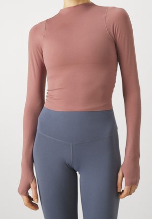 Haut moulant mauve à manches longues avec passages pour les pouces, associé à des leggings gris taille haute sur une personne debout, sur un fond uni.