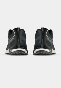 Melns un pelēks pārgājienu apavu modelis ar teksturētu auduma virsu, pastiprinātu papēdi un biezu gumijas zoli, kas ietver atstarojošas akcentus un saitēšanas sistēmu.