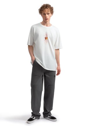 T-shirt en coton blanc avec un motif de plante, associé à un pantalon cargo gris ample avec plusieurs poches et des détails de couture.