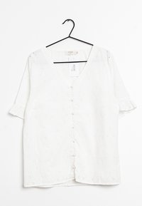 Blouse blanche à manches courtes avec un col en V, dotée d'un devant à boutons, d'un motif de broderie œillets et de manches retroussées. Accrochée à un crochet noir.