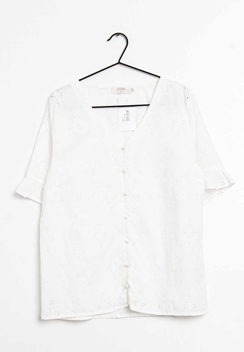 Blouse blanche à manches courtes avec un col en V, dotée d'un devant à boutons, d'un motif de broderie œillets et de manches retroussées. Accrochée à un crochet noir.