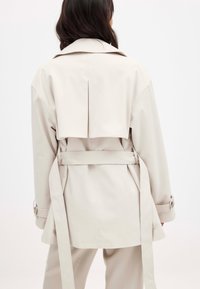 Šviesiai rudas trenčkotas su dideliu nugaroje esančiu ventiliaciniu skyriumi, diržu ant liemens ir užsegamais rankogaliais. Minkšta medžiaga su lygiu paviršiumi ir minimaliu dekoratyvumu.