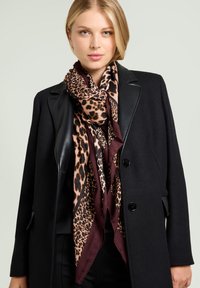 Seidenschal mit Leopardenmuster in Beige und Braun, mit dunkelburgunderfarbenen Akzenten, um den Hals getragen über einem schwarzen Mantel.