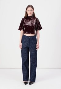 Burgundia sädelev crop top kõrge kaelusega, mis on kombineeritud tumedate siniste pükstega, millel on täppide muster. Modell seisab neutraalses taustas.