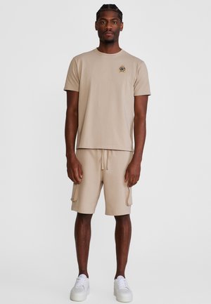 Mann steht und trägt ein beigefarbenes Kurzarm-T-Shirt mit Logo und passende Cargo-Shorts, kombiniert mit weißen Turnschuhen vor einem schlichten Hintergrund.