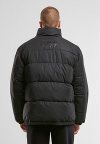 Sort puffer-jakke med høj krave, quiltet design, elastisk kant og et diskret logo på ryggen. Har brede polstrede sektioner.