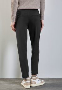 Pantalones negros ajustados de tela suave con dos bolsillos traseros y acentos laterales, combinados con zapatillas blancas y beige sobre una superficie gris.
