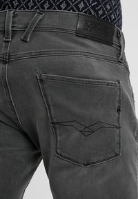 Šedé denimové džíny s zadním kapesním detailem s výraznou trojúhelníkovou výšivkou a černou koženou etiketu s nápisem "REPLAY" na pasu.