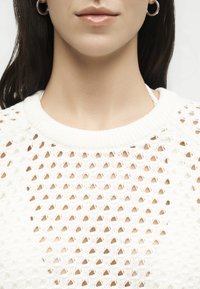 Γυναίκα που φοράει ασημένια κρίκους σκουλαρίκια και λευκό πλεκτό πουλόβερ με υφή και ανοιχτό πλέγμα.