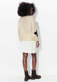 Pull en tricot beige avec un motif floral noir sur la manche, associé à une jupe en denim blanche et des bottines en daim marron.