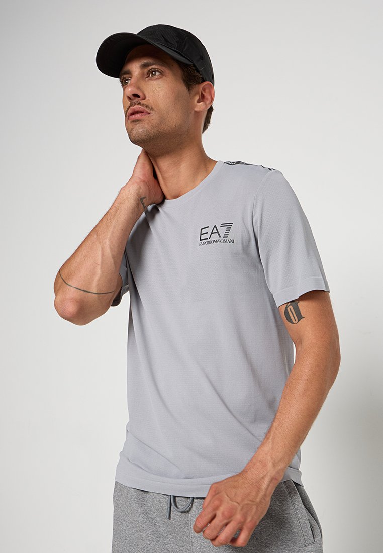 Světlé šedé tričko s krátkým rukávem a kulatým výstřihem, s černým logem na hrudi. K tričku jsou oblečeny šedé tepláky a černá čepice.