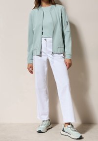 Kobieta w jasnozielonej kurtce zapinanej na zamek, z pasującą bluzką, białych prostych spodniach i jasnoniebieskich trampkach, na neutralnym tle.