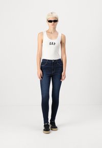 Fehér ujjatlan póló "GAP" logóval, sötétkék skinny farmernadrág, fekete napszemüveg és fekete sportcipő bézs talppal. Minimalista háttér.