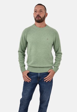 Grüner Strickpullover mit Rundhalsausschnitt und gerippten Bündchen; mit Logodetail; kombiniert mit dunkelblauen Jeans. Glatte Textur.