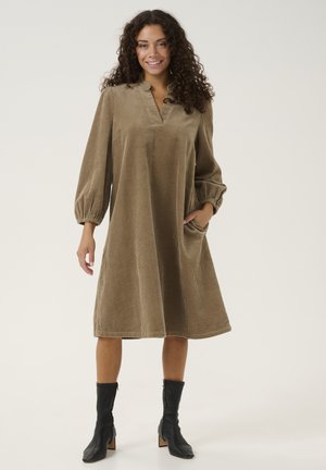 Robe en velours côtelé marron avec de longues manches bouffantes, un col en V et des poches latérales, atteignant le milieu du mollet. Assortie à des bottines noires.