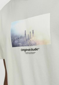 Camiseta de algodón gris claro con una impresión fotográfica de un paisaje urbano y el texto "Originals Studio" debajo en negro. Mangas cortas, cuello redondo.