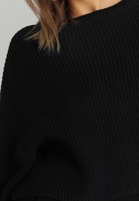 Maglione nero a maglia con una texture a coste, caratterizzato da un ampio scollo e una vestibilità morbida. Il tessuto appare morbido e spesso.