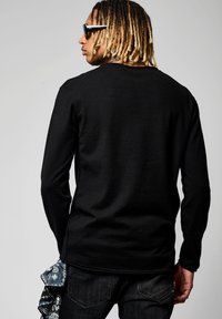 Maglietta nera a maniche lunghe con colletto girocollo e texture a costine, abbinata a jeans neri. Una bandana blu con un motivo è visibile in vita.