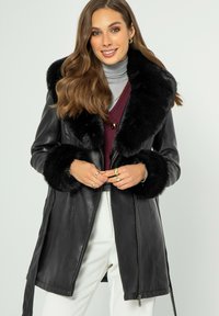 Giacca di pelle nera con un ampio colletto e polsini in pelliccia sintetica. Presenta una chiusura con zip e un design allungato, sovrapposta a un dolcevita grigio.
