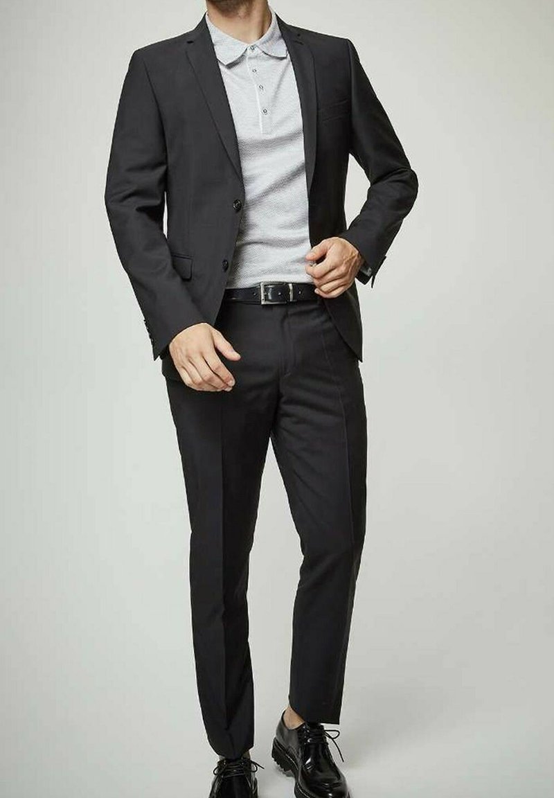 Costume noir sur mesure avec un polo gris en dessous. Comprend une veste à simple boutonnage, un pantalon slim et des chaussures noires brillantes.