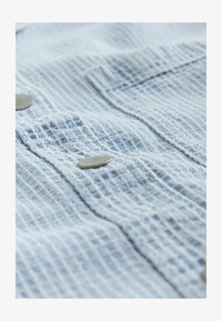 Tejido texturizado azul claro con un patrón de cuadrícula y botones de metal en dos bolsillos del pecho; apariencia suave y ligeramente arrugada.