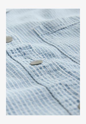 Jasnoniebieska, teksturowana tkanina w kratkę z metalowymi guzikami na dwóch kieszeniach na piersiach; miękka i lekko pomarszczona.