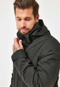 Man with beard adjusting collar of dark hooded winter jacket with visible sleeve logo against white background.