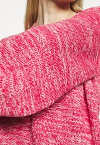 Chunky gebreide trui, roze met een lichter gemarmerd patroon, met geribbelde mouwen en een zachte, textuurachtige stof. Close-upbeeld benadrukt de ontwerpsdetails.
