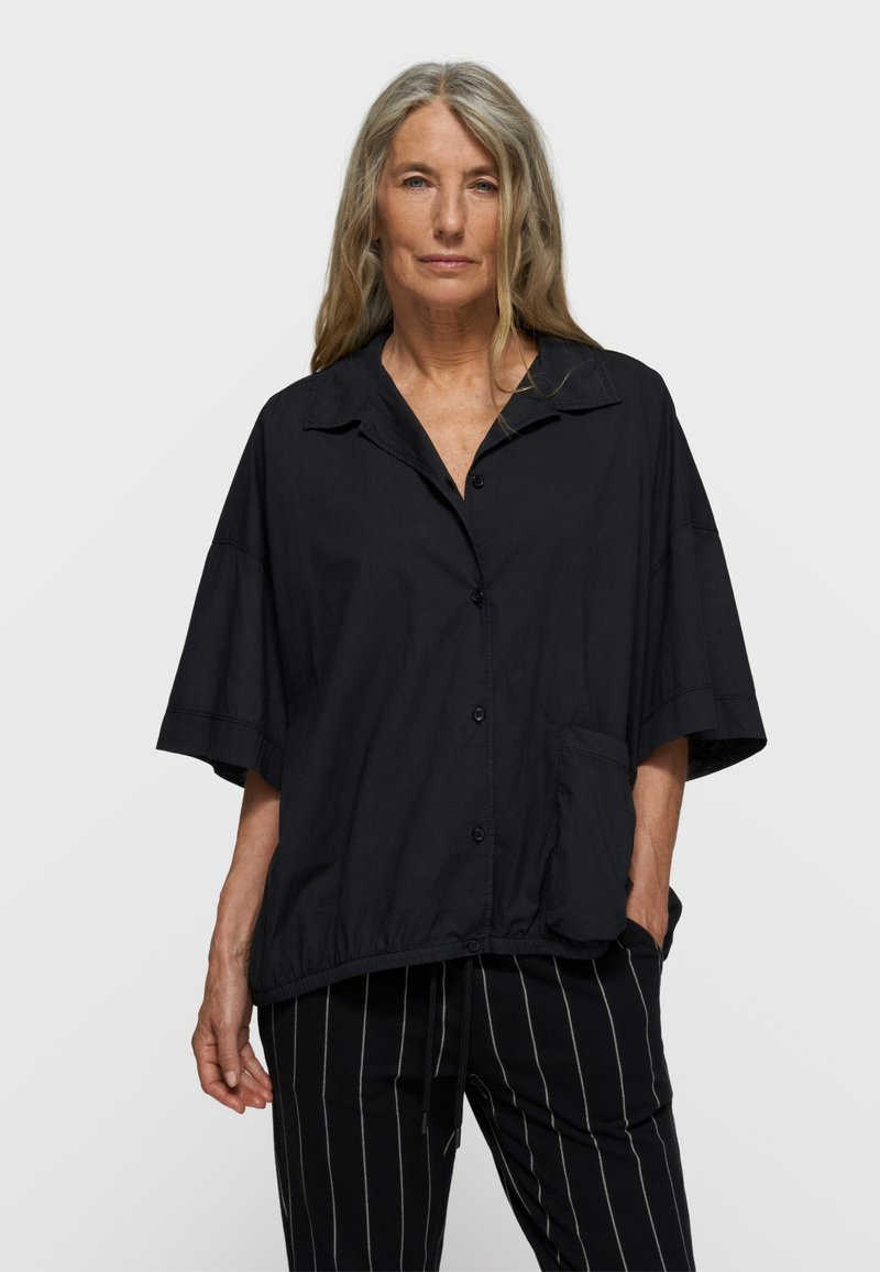 Chemise noire oversize à boutons avec col pointu, manches courtes et poche avant. Le matériau semble léger et lisse.