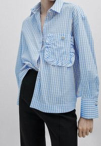 Camicia a righe blu e bianche con tasca arricciata, maniche lunghe e dettagli in bottoni argentati. Indossata con pantaloni neri.